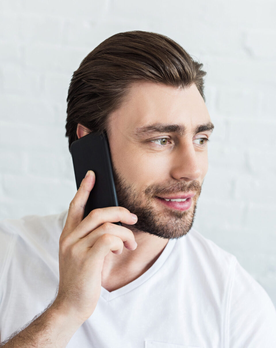 young smiling man talking on smartphone in front of white brick wall